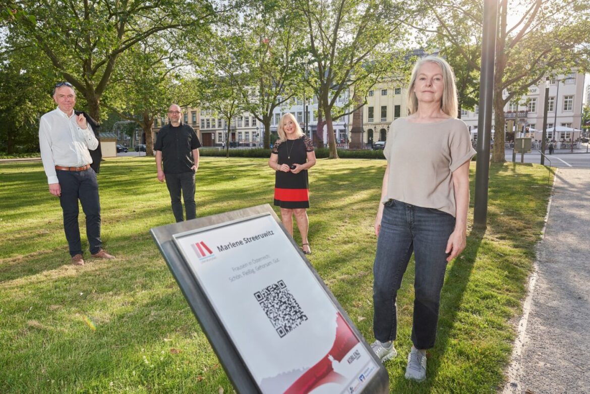 Vier Personen stehen auf einem Rasen neben einem Schild mit QR-Code.