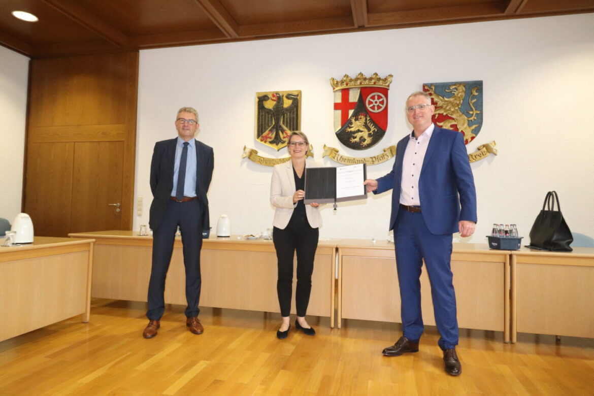 Drei Personen bei einer Urkundenübergabe in einem Konferenzraum.
