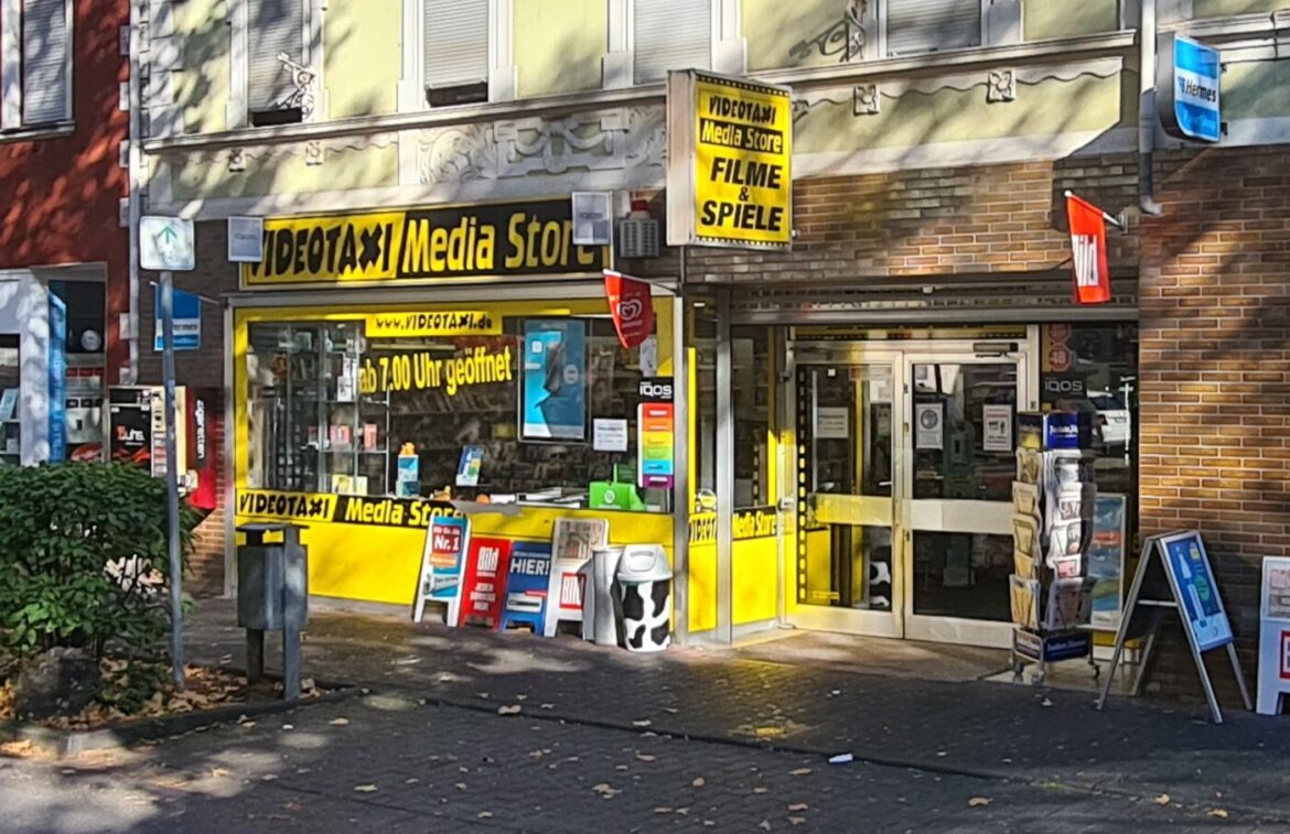 Eingang eines Media Stores mit gelbem Schild und Werbeplakaten.