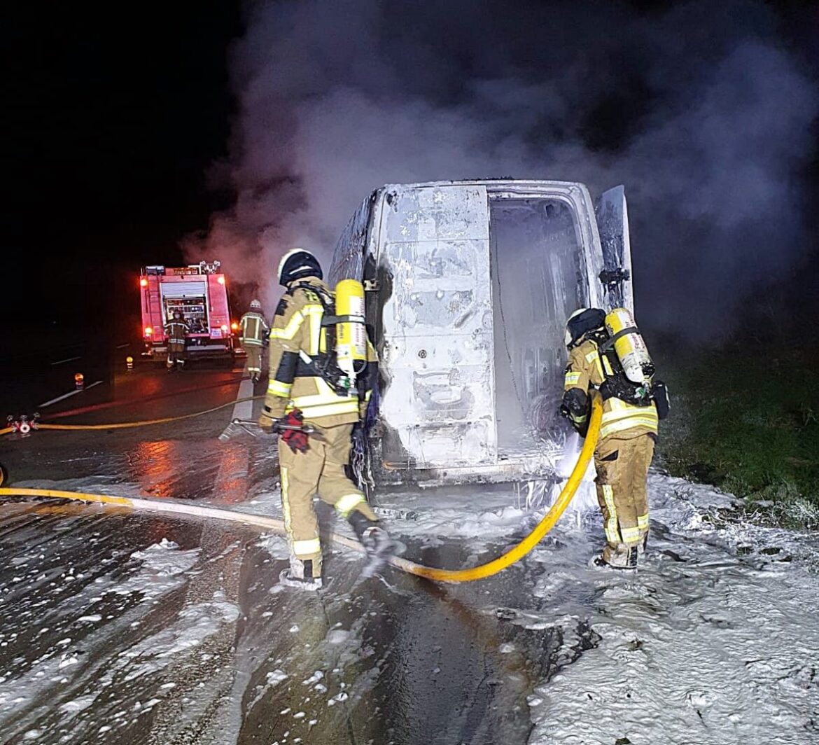 Feuerwehr löscht brennendes Fahrzeug auf Straße bei Nacht.