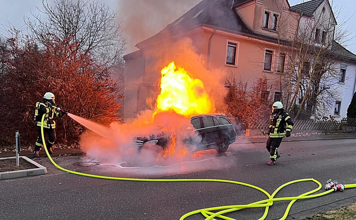 Feuerwehr löscht brennendes Auto vor einem Haus.