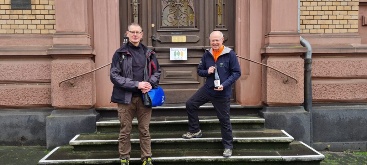 Zwei Männer stehen vor einem historischen Gebäude mit einer Flasche und einer Tasche.