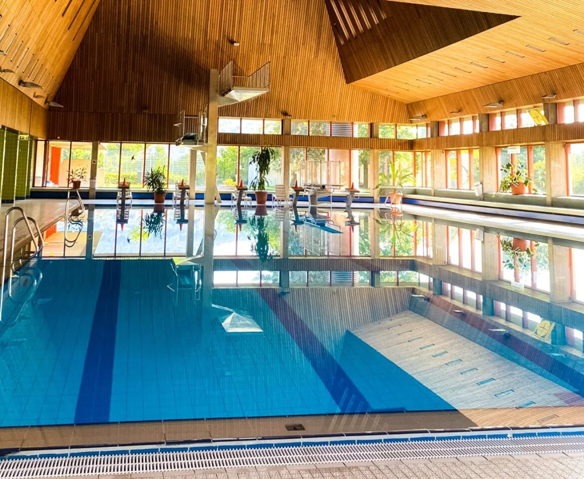 Innenansicht eines Hallenbads mit blauem Wasser und Holzdecke.