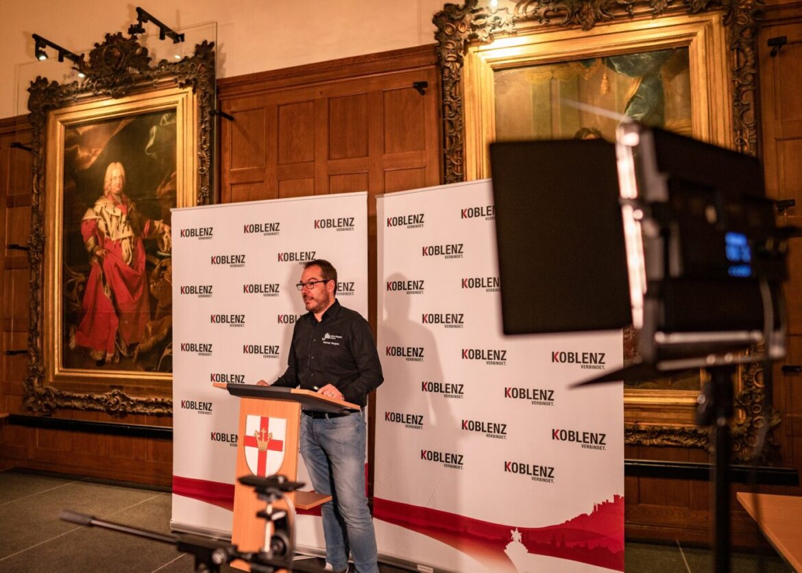 Mann hält Vortrag vor Koblenz-Banner in historischem Raum.