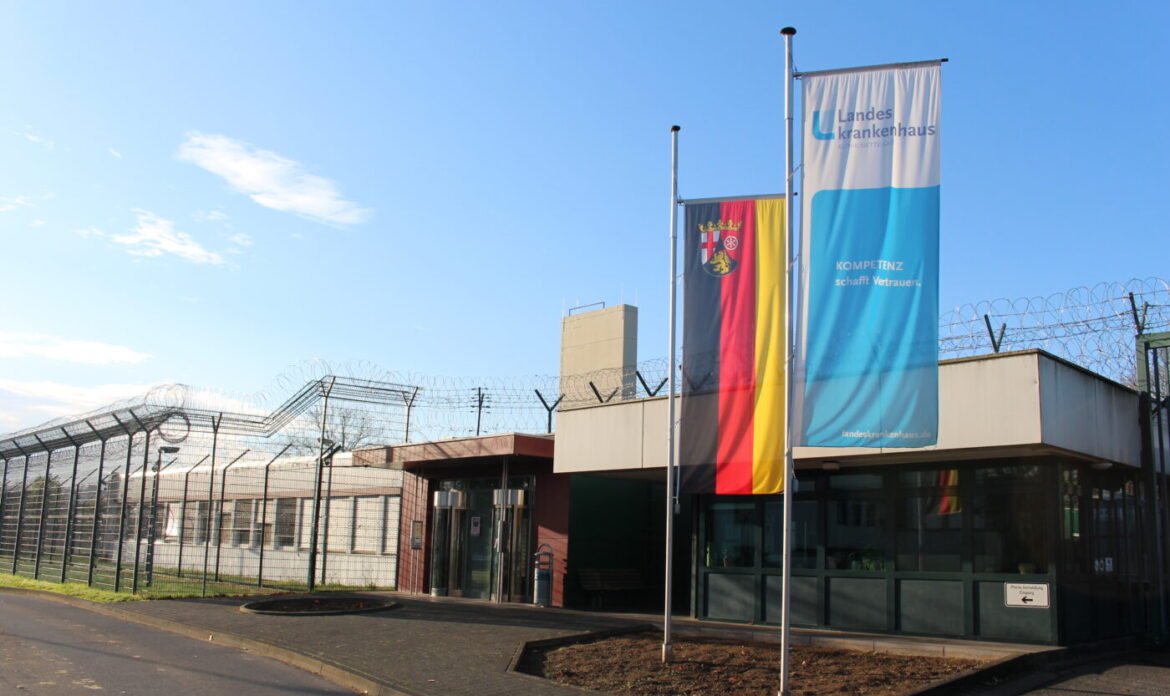 Eingang eines Landeskrankenhauses mit Zaun und Flaggen im Vordergrund, strahlend blauer Himmel.