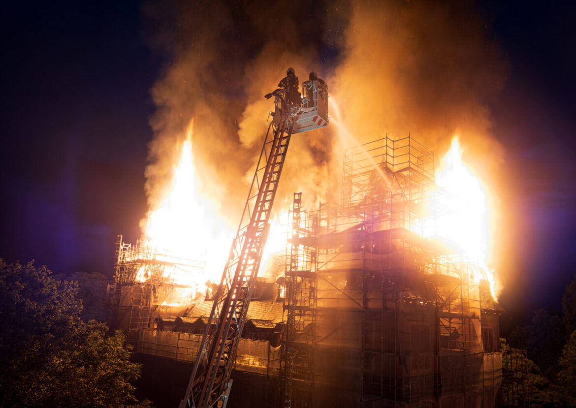 Ein brennendes Gebäude mit Flammen und Rauch, während Feuerwehrleute mit einer Drehleiter löschen.