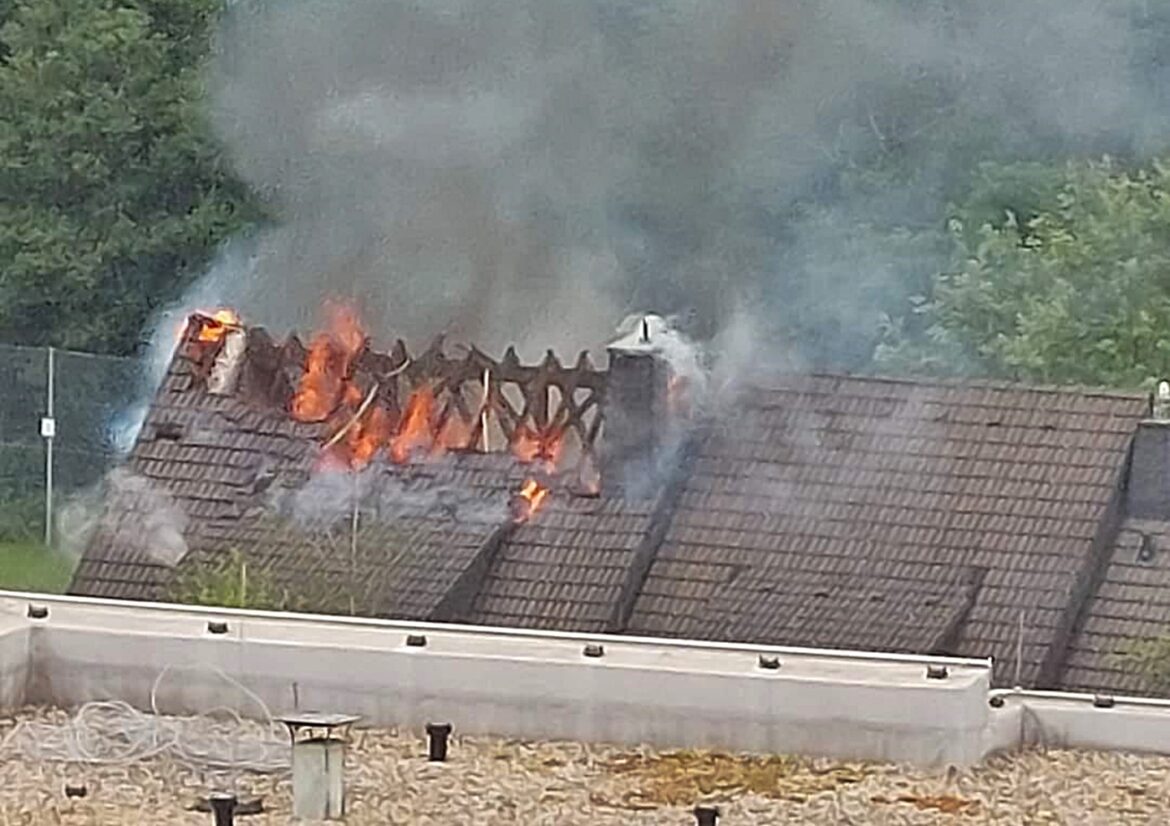 Dach eines Hauses brennt, Flammen und Rauch steigen auf. Grüne Bäume im Hintergrund.