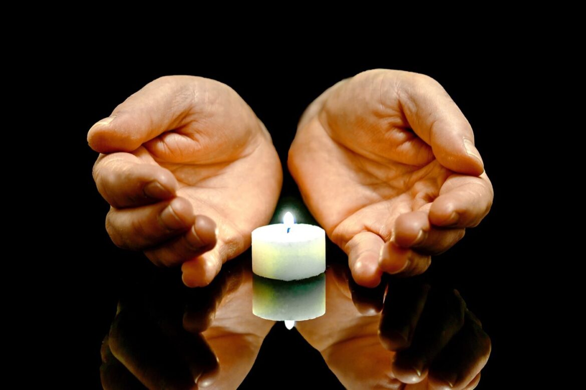 Zwei geöffnete Hände halten eine kleine Kerze auf einer reflektierenden Oberfläche, vor schwarzem Hintergrund.