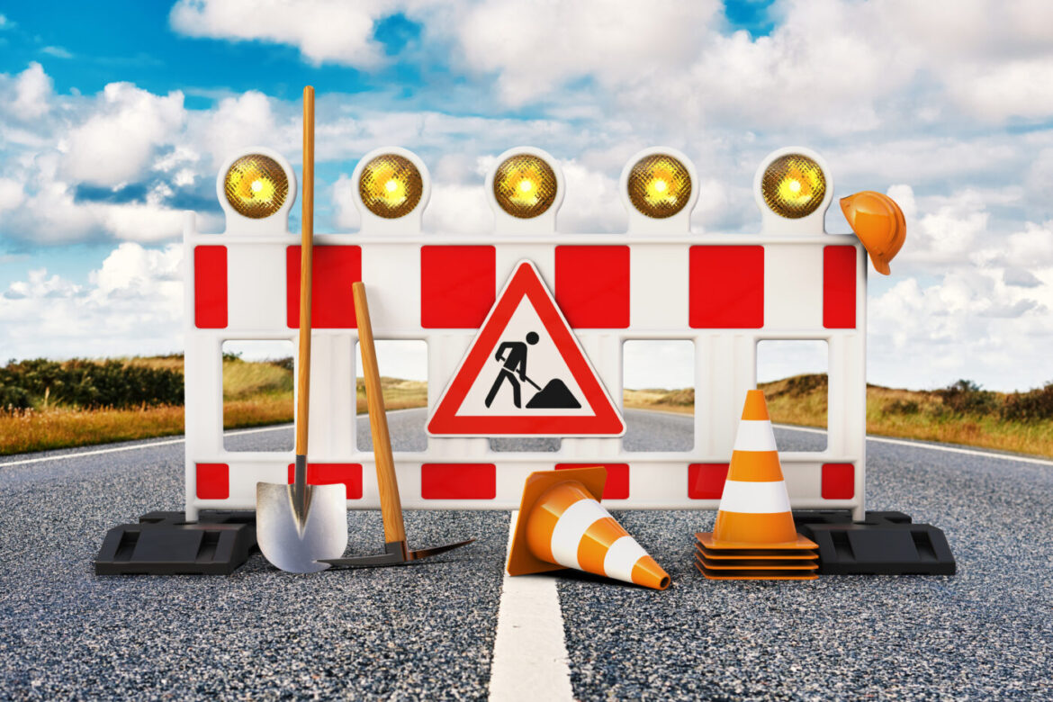 Baustellenabsperrung mit Warnlichtern, Schaufel, Pickel, Bauhelm und Verkehrshütchen auf einer Straße.