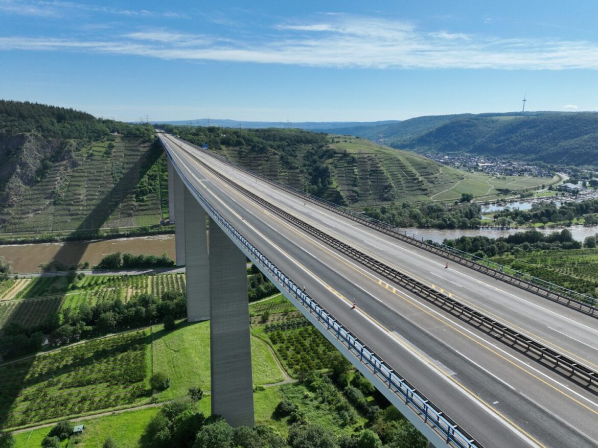Hochbrücke mit Aussicht auf Weinberge und Fluss im feuchten, grünen Tal unter blauem Himmel.