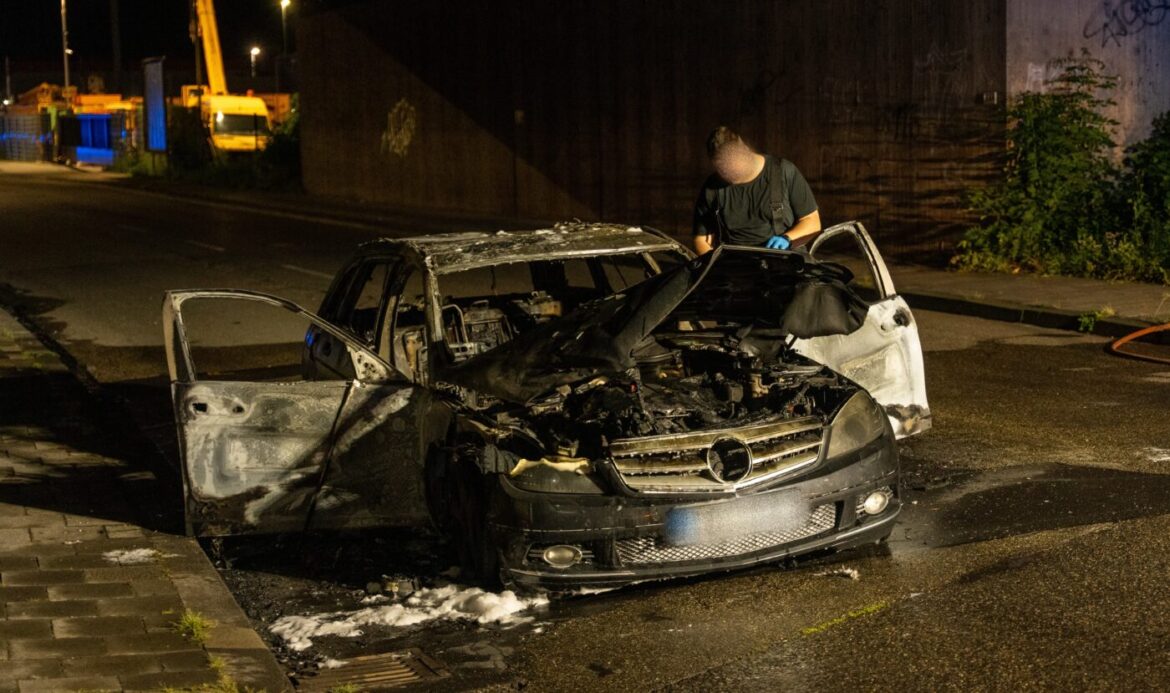Ein ausgebranntes Auto auf der Straße, ein Mann untersucht den Motorraum.