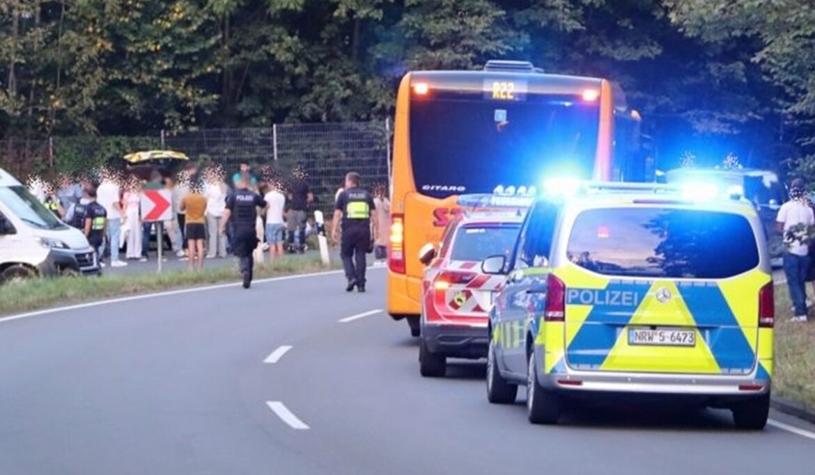 Polizeifahrzeuge stehen an einer Landstraße, eine Menschenmenge ist im Hintergrund zu sehen.