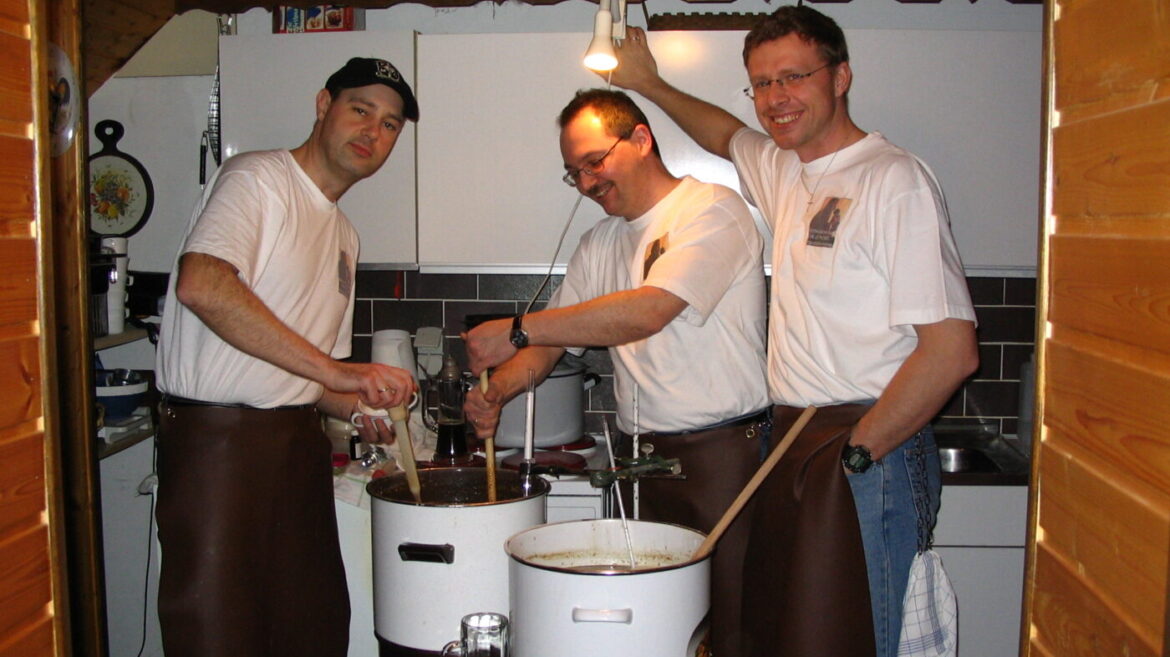 Drei Männer in Schürzen brauen gemeinsam Bier in einer Küche. Töpfe und Utensilien sind sichtbar.