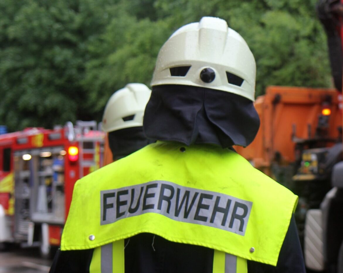 Feuerwehrmann mit gelber Warnjacke und Helm, im Hintergrund Feuerwehrauto und grüne Bäume.