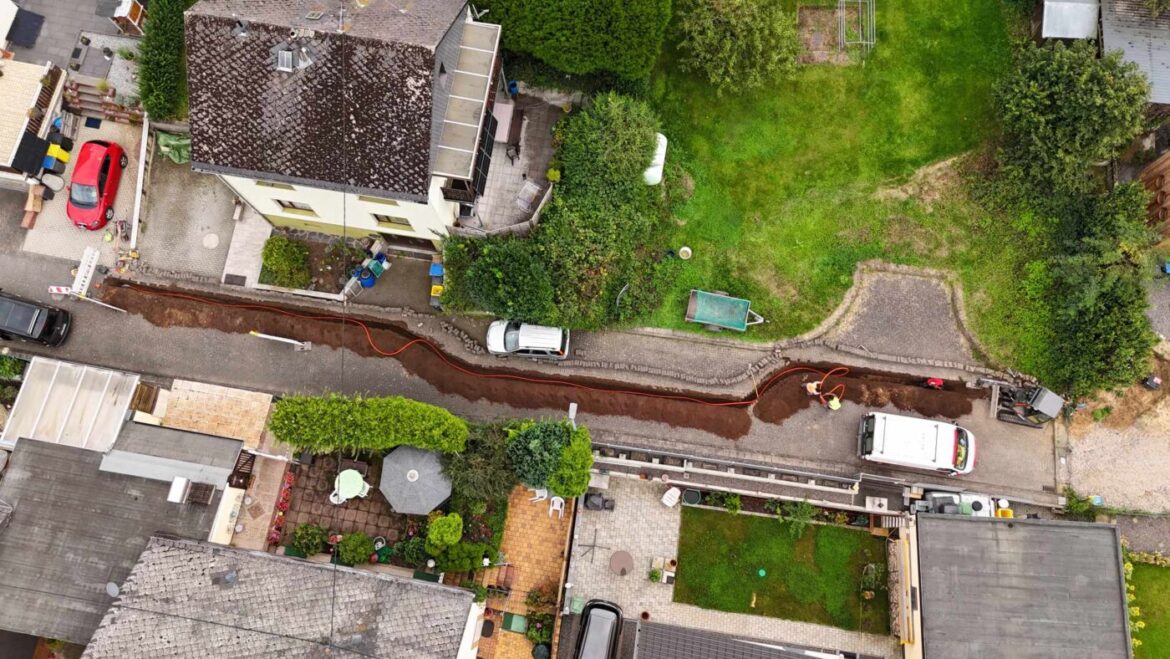 Luftaufnahme einer Straße mit Baustelle, Erdarbeiten und zwei Fahrzeugen in Wohngegend.