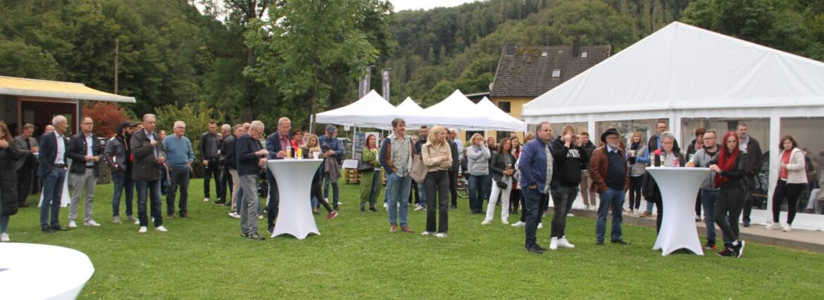 Eine Gruppe von Menschen steht auf einer Wiese, umgeben von Zelten und Bäumen, bei einem Event.