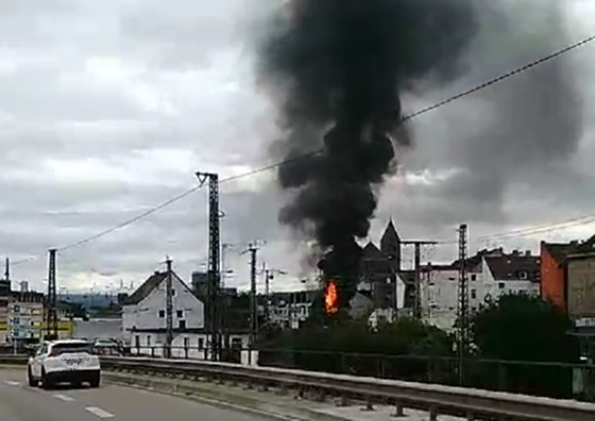 Rauch und Flammen steigen aus einem Gebäude in einer städtischen Umgebung auf, während ein Auto vorbeifährt.