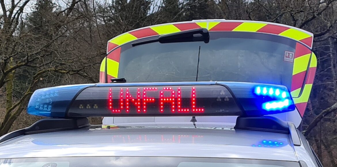 Blaulicht auf einem Einsatzfahrzeug mit einer Anzeige, die "UNFALL" zeigt, im Hintergrund ein Fahrzeug mit reflektierenden Streifen.