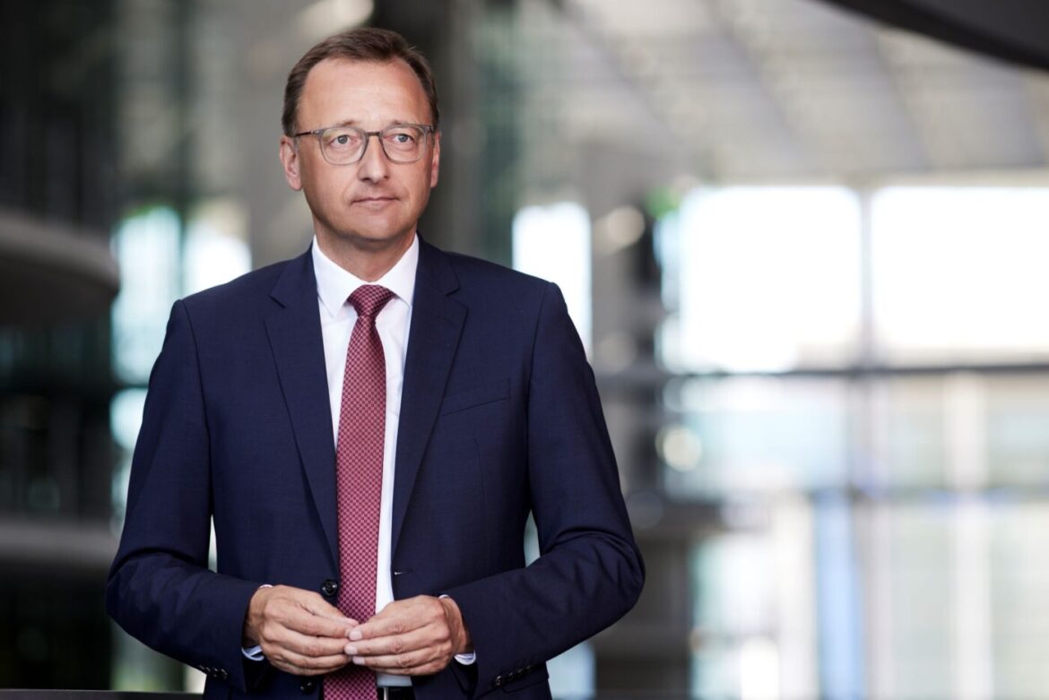 Mann in Anzug mit Krawatte steht in einem modernen Innenraum und schaut direkt in die Kamera.