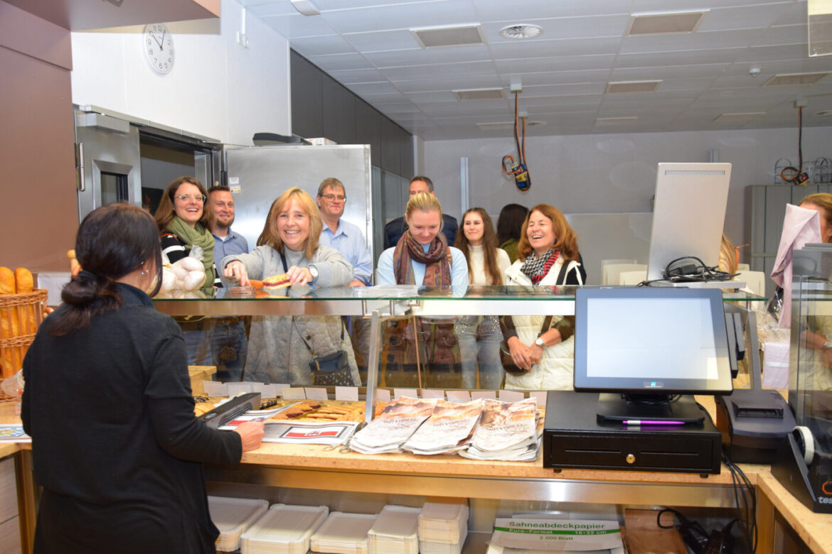 Personen stehen lächelnd an einem Bäckereitresen, Verkäuferin bedient sie.