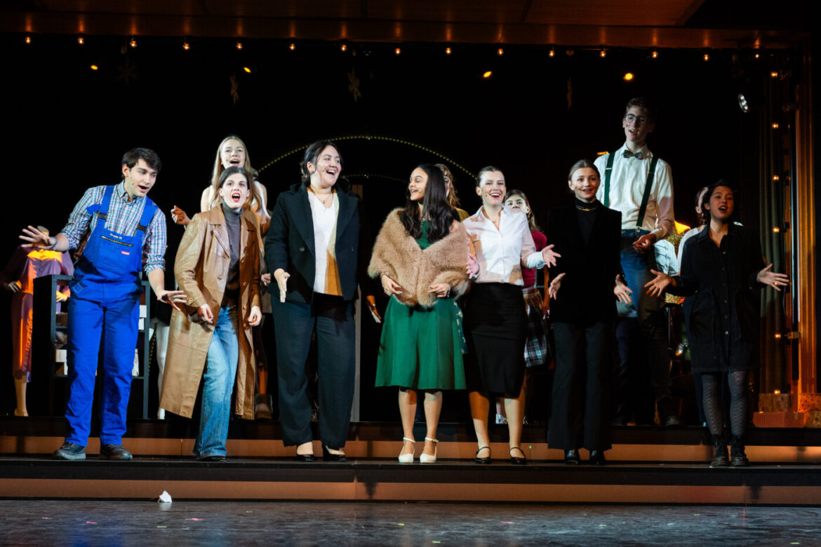 Gruppe junger Menschen in unterschiedlicher Kleidung auf einer Bühne singend und tanzend, Hintergrund beleuchtet.