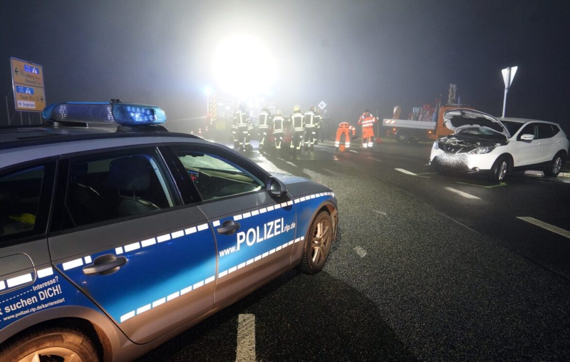 Polizeiauto im Vordergrund, Autounfall bei Nacht, Rettungskräfte und beschädigtes weißes Auto im Hintergrund.