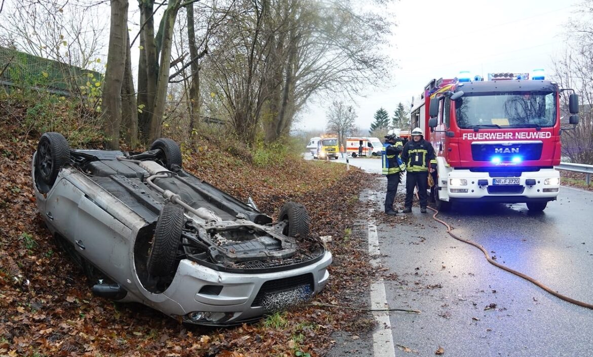 Verunglücktes Auto auf dem Dach, Feuerwehrwagen und Einsatzkräfte an regennasser Landstraße.