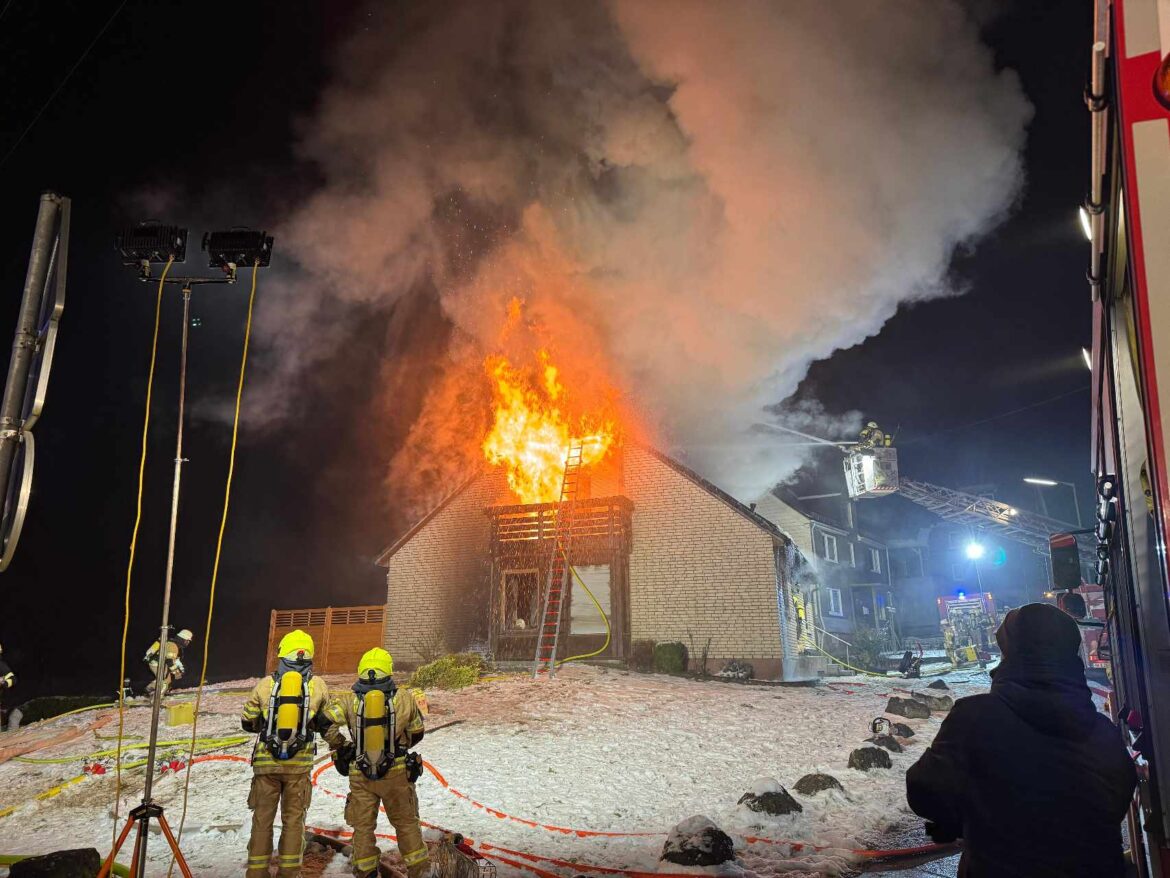 Brennendes Haus bei Nacht, Feuerwehrleute mit Atemschutzgeräten, Einsatzfahrzeuge und Leiter im Einsatz.