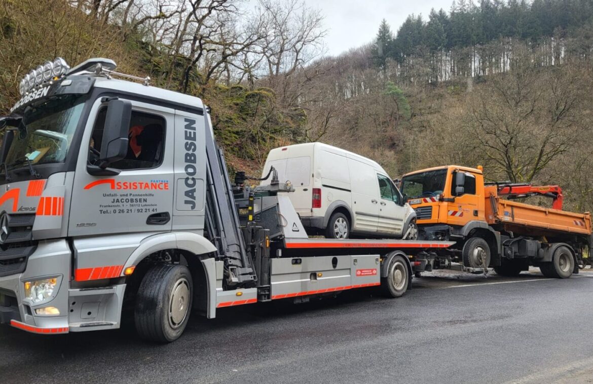 Abschleppwagen transportiert weißen Lieferwagen, orangefarbener LKW auf bewaldeter Straße im Hintergrund.