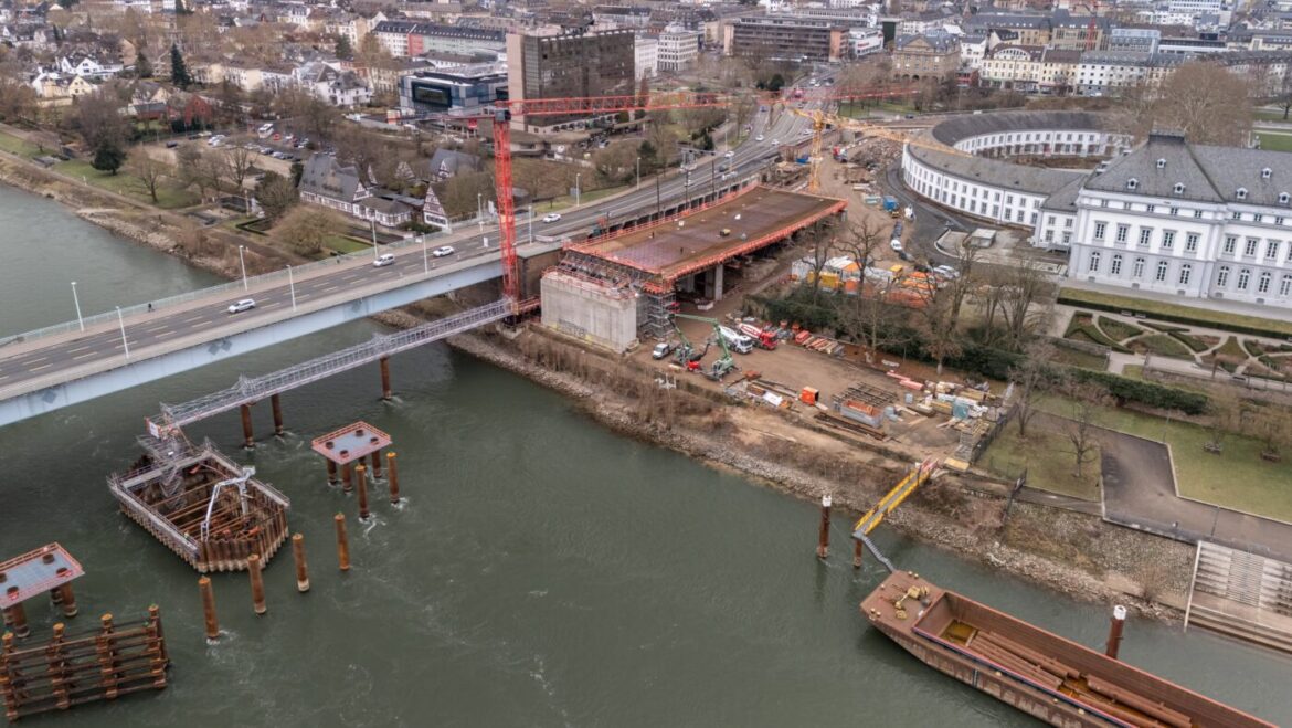 Luftaufnahme einer Brückenbaustelle über einem Fluss, mit umliegenden Gebäuden und Kränen.