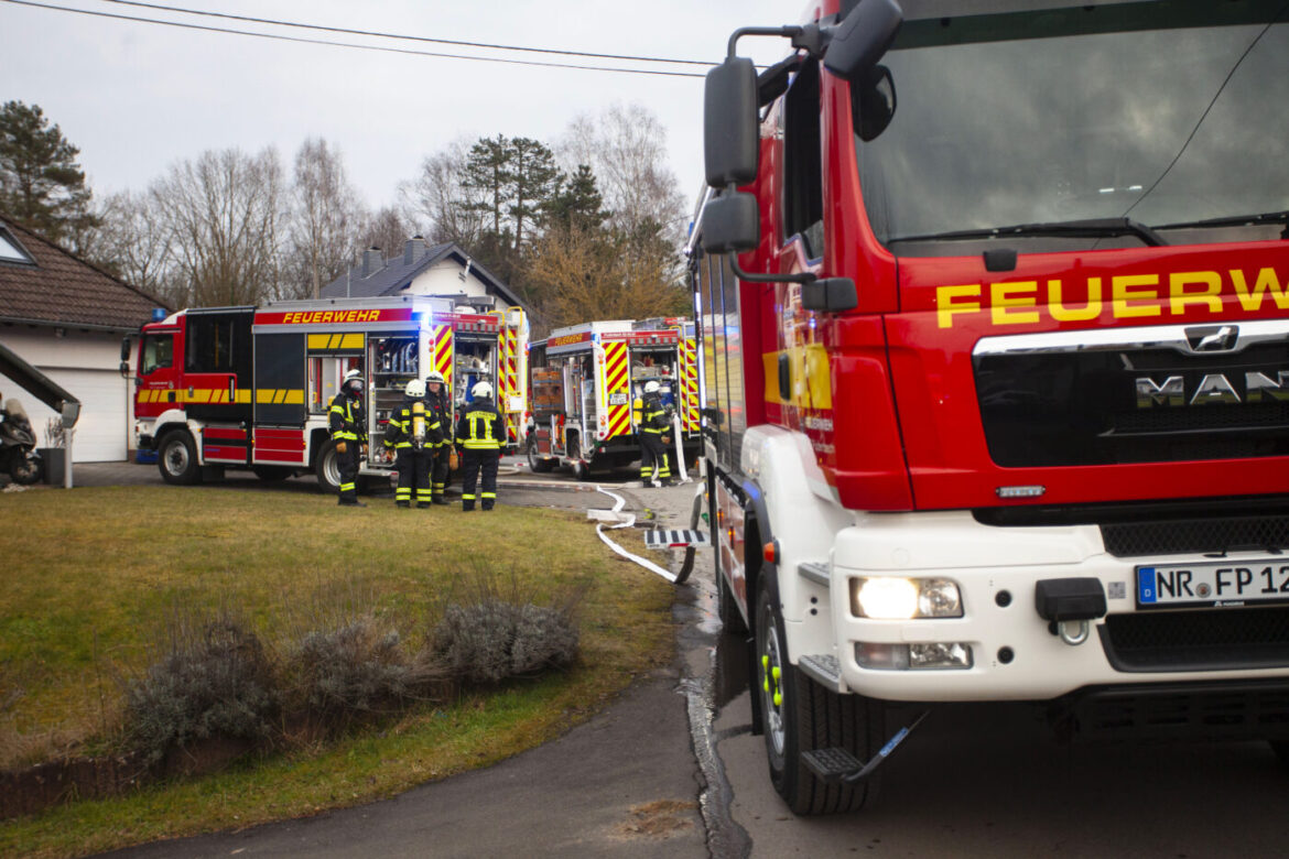 Feuerwehrfahrzeuge und Feuerwehrleute in Einsatzkleidung vor Wohnhaus mit Schläuchen auf der Einfahrt.