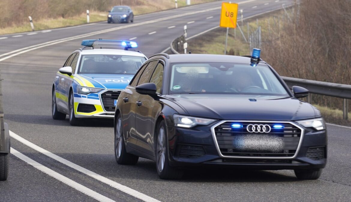 Schwarzer Audi mit Blaulicht auf Straße, gefolgt von Polizeiauto mit eingeschaltetem Blaulicht.