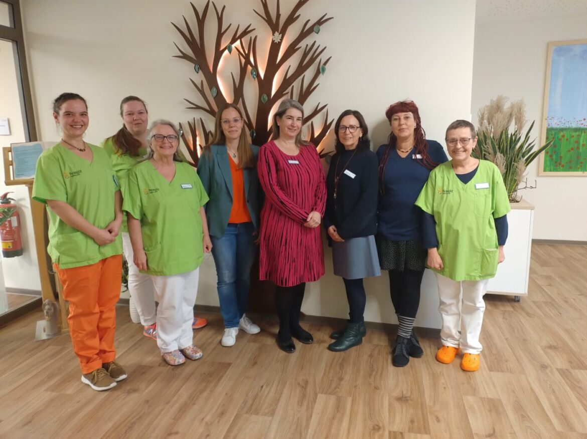 Neun Personen stehen lächelnd in einem Raum; einige tragen grüne Arbeitskleidung, im Hintergrund Baum-Dekoration.
