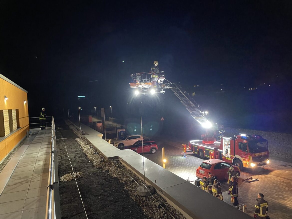 Feuerwehr im Nachteinsatz, Leiterfahrzeug mit Licht, Personen auf Dach und Straße, gelbes Gebäude links.