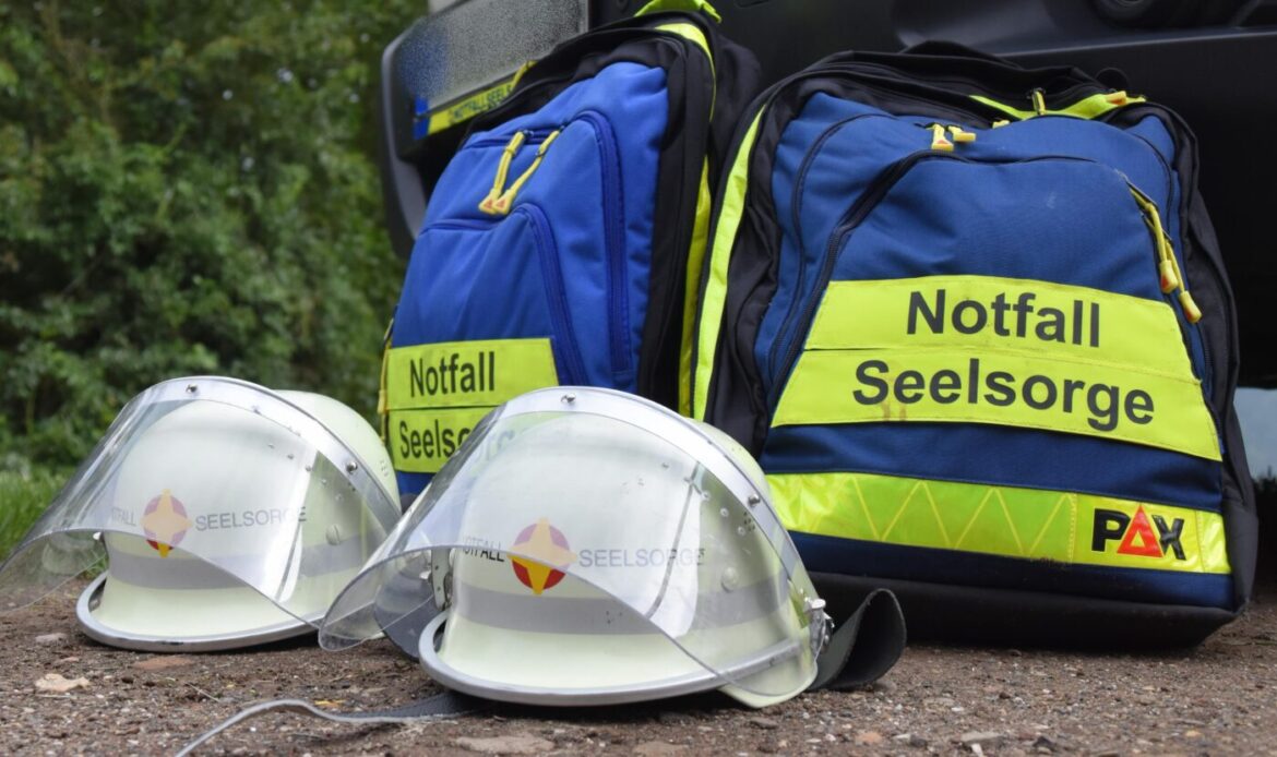 Zwei blaue Rucksäcke und weiße Helme mit Notfallseelsorge-Beschriftung auf einem Boden im Freien.