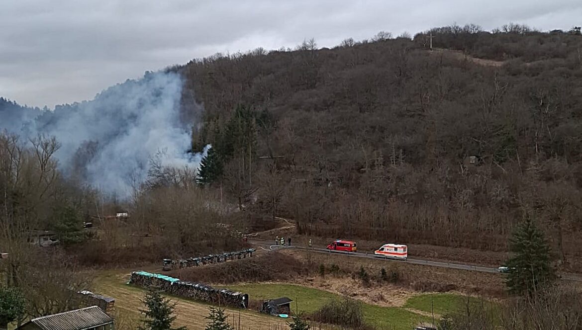 Rauch über bewaldetem Hügel, Feuerwehrfahrzeuge und Rettungswagen auf Straße im Vordergrund.