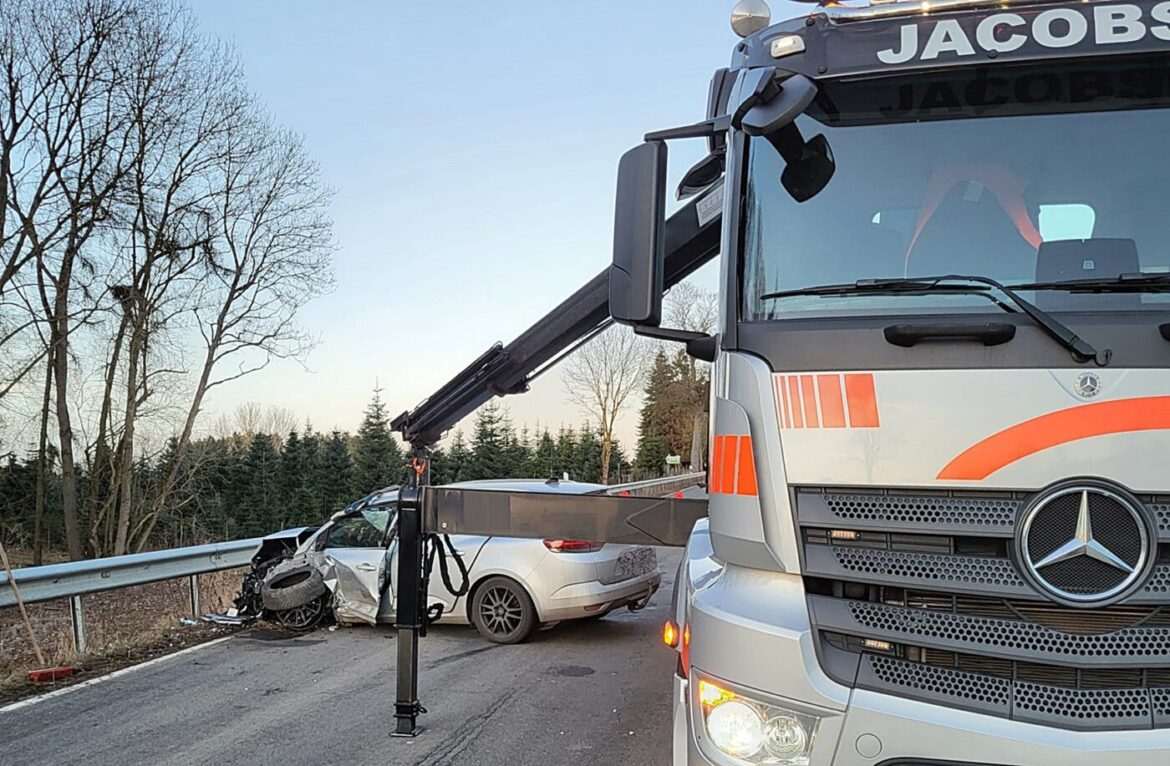 Abschleppwagen hebt beschädigtes Auto nahe Leitplanke, Bäume im Hintergrund, winterliche Umgebung.