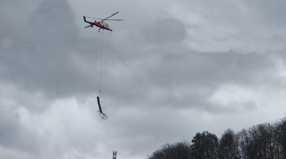Hubschrauber transportiert Baumstamm über bewölktem Himmel, Wald im Hintergrund.