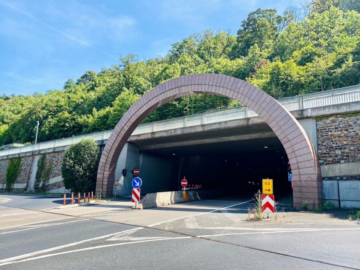 Straßentunnel mit gemauerter Bogenstruktur, umgeben von Bäumen, mit Verkehrsschildern im Vordergrund.