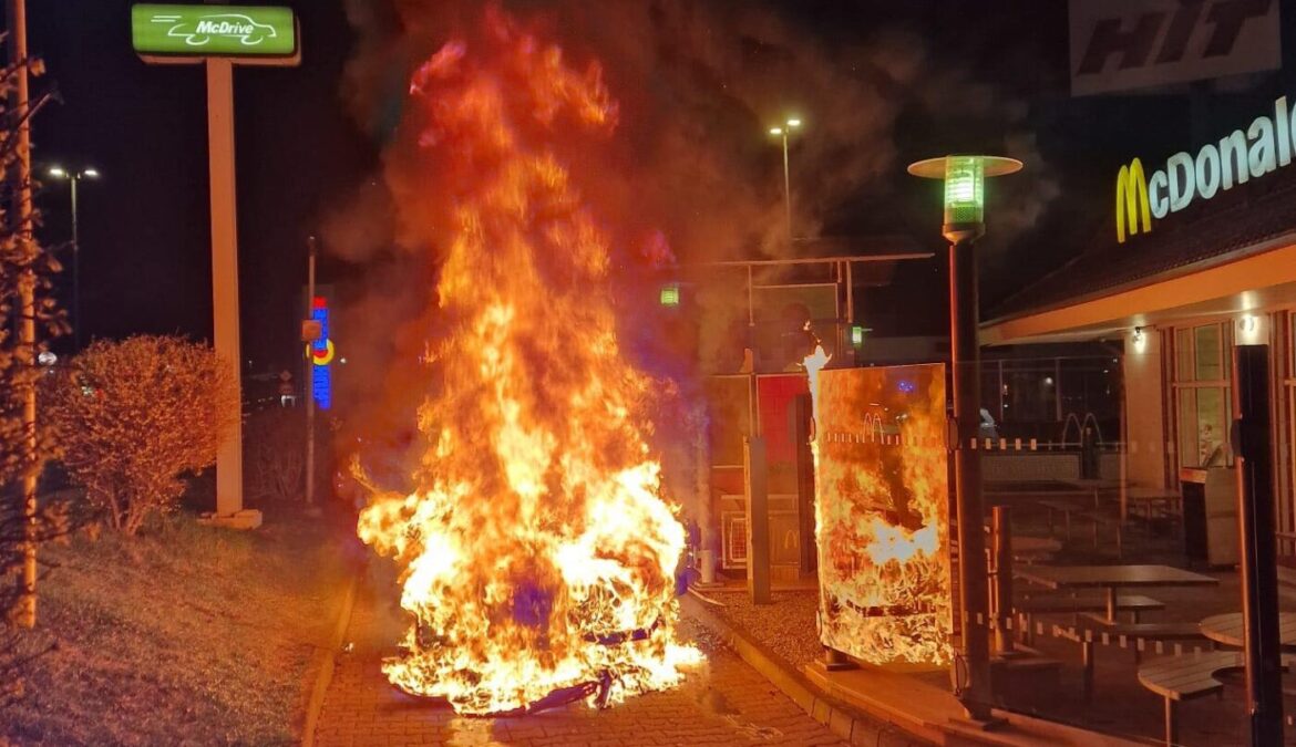 Auto brennt nachts vor McDonald's-Filiale, Flammen und Rauch sichtbar, beleuchtetes Schild im Hintergrund.