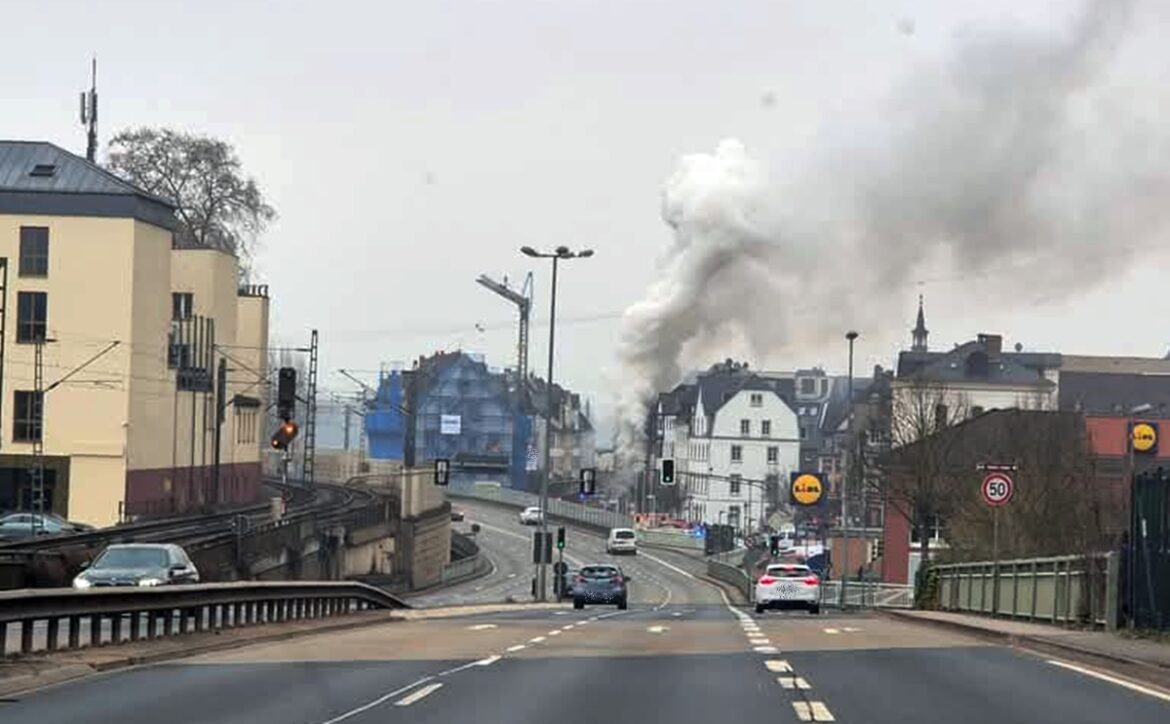 Straße mit Autos, Rauch steigt hinter Gebäuden auf, Verkehrsampeln und Baustelle im Hintergrund.