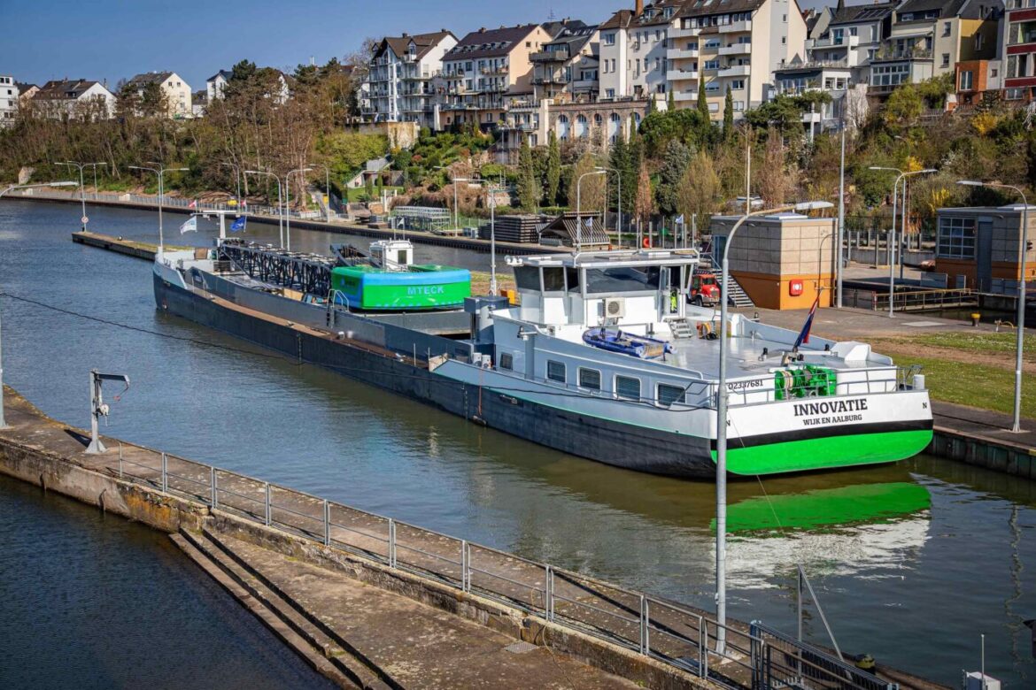 Frachtschiff auf einem Fluss oder Kanal, umgeben von Wohnhäusern und grüner Vegetation im Hintergrund.