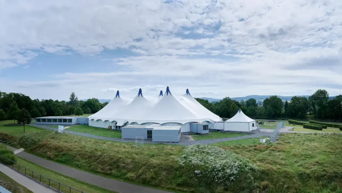 Großes weißes Zelt mit blauen Spitzen in grüner Landschaft und bewölktem Himmel.