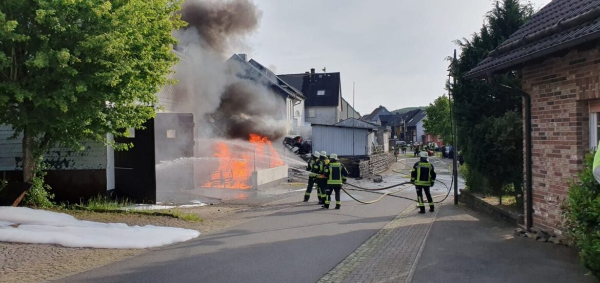 Feuerwehrleute löschen Feuer in einer Wohnstraße, schwarzer Rauch steigt auf, umliegende Gebäude sichtbar.