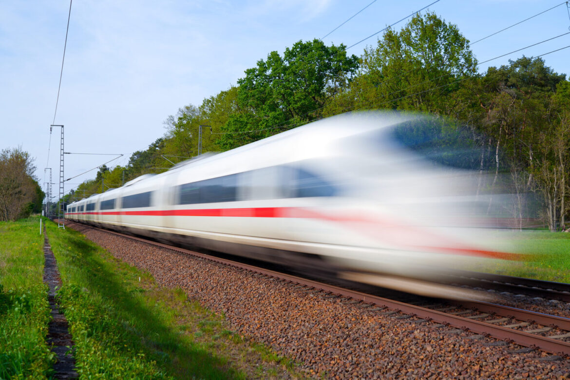 Schnellzug fährt bei Tageslicht durch grüne Landschaft, Bewegung unscharf dargestellt.