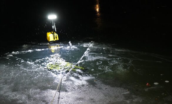 56aktuell_BWTW2EZe Gelbes Tauchlicht beleuchtet gefrorenen Teich, rechteckiges Loch im Eis, Regentropfen sichtbar.