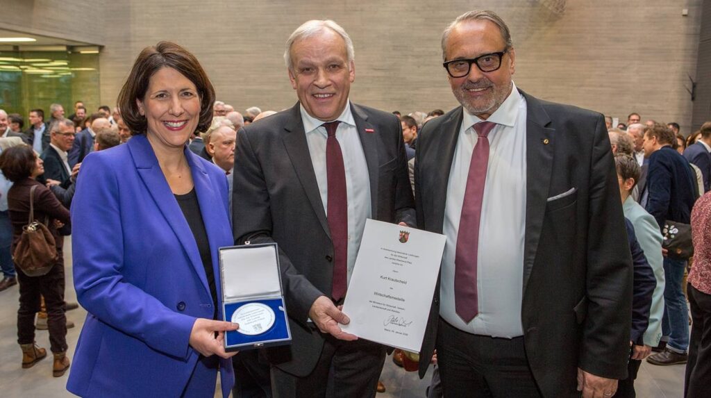 Drei Personen bei einer Preisverleihung mit Urkunde und Medaille.